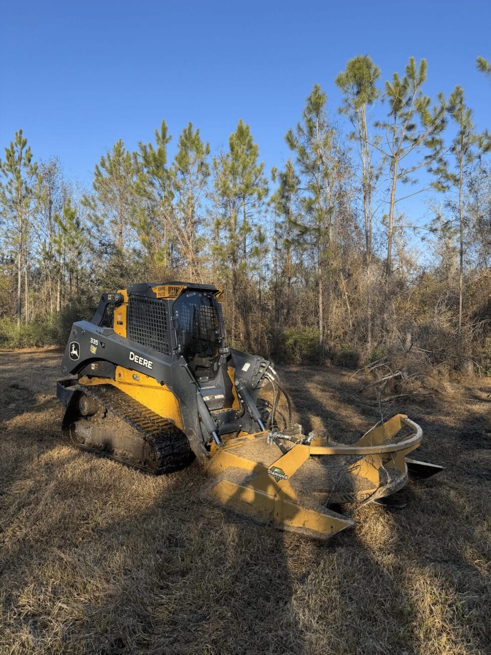 A yellow and black skid steer loader with a forestry attachment is parked on grassy terrain, surrounded by trees under a clear blue sky.